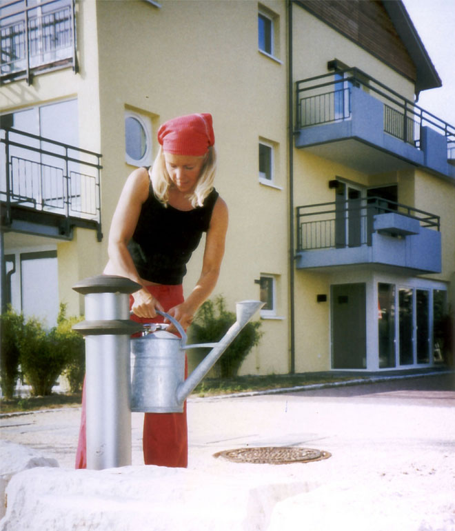 Eine Frau befüllt eine Gießkanne vor einem Mehrfamilienhaus